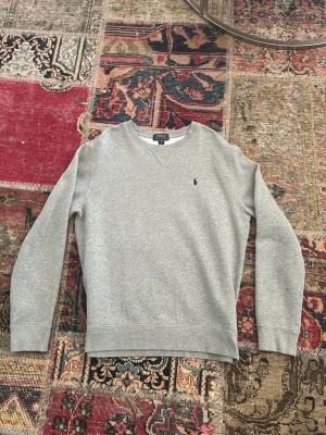 Är som en S, är XL i barn - Klassisk grå sweatshirt från Polo Ralph Lauren med rund halsringning och diskret broderad logga på bröstet. Tröjan har ribbade muddar vid ärmslut och nederkant, perfekt för en avslappnad och stilren look. Det är storlek XL i barn så den fungerar som en S i herr.