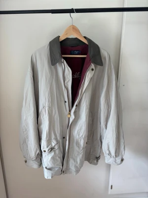 Beige rock från Gant med kontrastkrage - Säljer en beige rock från Gant, modell The Slugger, med mörkgrå kontrastkrage och vinrött innerfoder. Rocken har stora fickor framtill, knapp- och dragkedjestängning samt avslappnad passform. Perfekt för dig som gillar klassisk och tidlös stil.