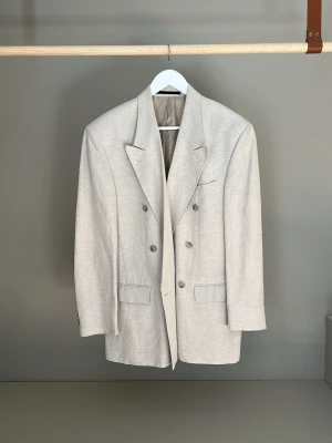 Beige dubbelknäppt linnekavaj från Tiger of Sweden - Snygg beige kavaj från Tiger med dubbelknäppning och breda slag. Kavajen har fyra knappar framtill, två fickor med lock och en bröstficka. Lite boxiga axlar och rak form. Materialet är en blandning av linne och viscose för en avslappnad men stilren look.