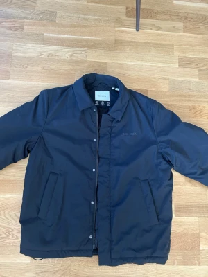 Svart vindjacka från Les Deux - Stilren svart vindjacka från Les Deux med klassisk krage och dragkedja samt tryckknappar framtill. Två snedställda fickor och diskret logga på bröstet. Jackan är tillverkad i ett vattenavvisande material och har en minimalistisk look.