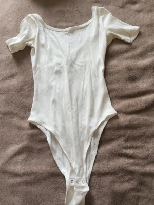 Vit ribbad body med kort ärm - Säljer en enkel och stilren vit body med korta ärmar och rundad halsringning. Använd 1 gång.
