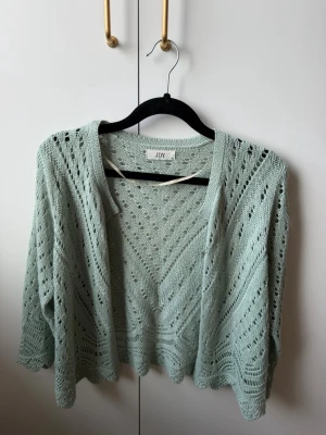Ljusgrön stickad cardigan från JDY - Snygg ljusgrön stickad cardigan från JDY med öppet hålmönster och lös passform. Tröjan har trekvartsärmar och vågig kant nertill. Perfekt att slänga över en topp för en chill look.