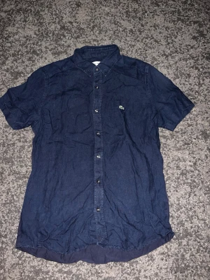 Mörkblå kortärmad skjorta - Säljer en mörkblå skjorta med korta ärmar och klassisk krage. Skjortan har knappar framtill och en liten broderad logga på bröstet. Perfekt till jeans eller chinos för en clean och enkel stil.