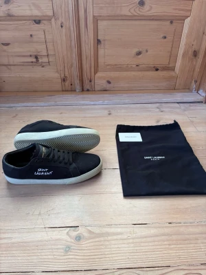Svarta Saint Laurent sneakers - Snygga svarta sneakers från Saint Laurent med vit sula och gulddetaljer på plösen. Skorna har lågt skaft och klassisk snörning. Materialet är canvas med Saint Laurent-logga broderad på sidan. Perfekta för en stilren och trendig look.