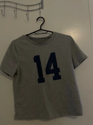 Grå t-shirt med blått tryck - Snygg grå t-shirt med mörkblått tryck av siffran 14 på framsidan. Klassisk rund halsringning och korta ärmar. Perfekt för en avslappnad och sportig stil.