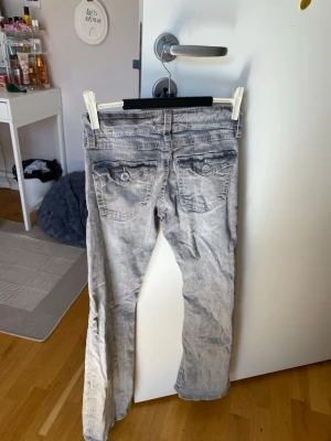 Grå  bootcut jeans - Säljer ett par grå tvättade jeans med bootcut passform och låg midja. Jeansen har fem fickor, knappgylf och slitna detaljer för en cool look. Perfekta för dig som gillar en avslappnad och trendig stil.