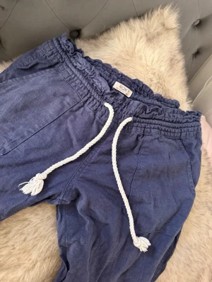 Blå linnebyxor från Roxy - Snygga trendiga blå linnebyxor från roxy med resår i midjan och vit snörning i repstil. Byxorna har två sidofickor fram och två bakfickor. Perfekta för en avslappnad och trendig look. Tillverkade i ett luftigt linnematerial. De är upsprätta i benen längst ner för att de var korta i storleken och jag ville ha dem längre. Storlek S. De är väldigt lågmidjade och raka ben.