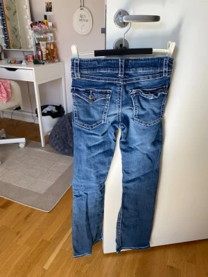 Blå bootcut jeans med kontrastsömmar - Säljer ett par blå bootcut jeans med vita kontrastsömmar och snygga detaljer på fickorna. Jeansen har låg midja, fem fickor och knappgylf. Perfekta för en avslappnad och trendig look.