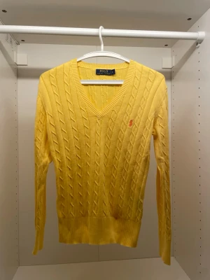 Gul kabelstickad tröja Polo Ralph Lauren - Snygg gul kabelstickad tröja från Polo Ralph Lauren med V-ringning och lång ärm. Tröjan har ribbade muddar och den klassiska lilla loggan broderad på bröstet. Perfekt för dig som vill ha en färgklick i garderoben.