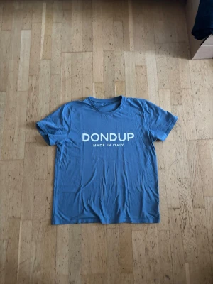 Dondup T-shirt - Säljer en blå t-shirt från Dondup med vit text 'DONDUP MADE IN ITALY' på bröstet. Klassisk rund hals och korta ärmar. Perfekt för dig som gillar enkel och stilren design.