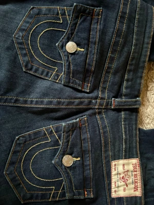 Mörkblå True Religion jeans - Säljer ett par mörkblå jeans från True Religion med ikoniska kontrastsömmar och stora fickor bak med lock och silverfärgade knappar. Klassisk femficksmodell och patch med logga bak i midjan. Snygg passform och slitstarkt denimtyg.