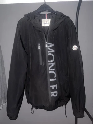 Svart vindjacka från Moncler - Snygg svart vindjacka från Moncler med stor logga längs dragkedjan och Moncler-märke på ärmen. Jackan har huva med dragsko, bröstficka med dragkedja och justerbar nederkant. Perfekt för dig som vill ha en stilren och sportig look.