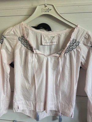 Ljusrosa Odd Molly blus med spets - Supersöt ljusrosa blus från Odd Molly med grå spetsdetaljer vid ringningen och axlarna. Blusen har långa ärmar och knytband framtill med tofsar. Tillverkad i mjukt bomullstyg och har en avslappnad, bohemisk stil.