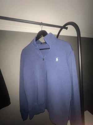 Blå quarter zip från Polo Ralph Lauren - Säljer en blå quarter zip sweatshirt från Polo Ralph Lauren med klassisk vit logga på bröstet. Tröjan har hög krage, dragkedja upptill och ribbade muddar vid ärmar och nederkant. Perfekt för en avslappnad och stilren look.