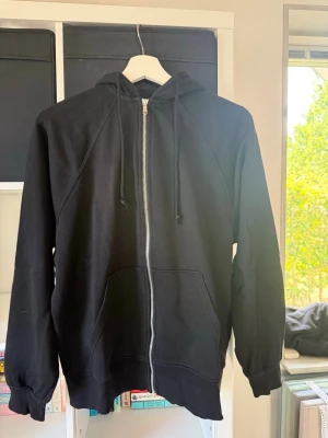 Svart zip hoodie med fickor - En klassisk svart hoodie med dragkedja framtill, huva med dragsnören och två stora fickor. 