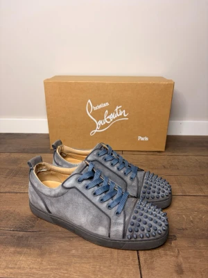  Christian Louboutin  - Unika grå sneakers från Christian Louboutin med blå skosnören och coola nitar på tån. Skorna har en låg profil och är tillverkade i mockaliknande material. Perfekta för dig som vill sticka ut med en edgy och exklusiv stil. Storlek 41. Skorna är 100% autentiska ✅