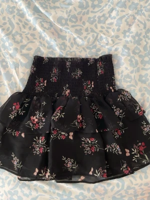 Svart blommig volangkjol - Söt svart kortkjol med blommigt mönster och bred smock i midjan. Kjolen har flera lager volanger som ger en luftig och lekfull känsla. Perfekt för dig som gillar romantisk stil och vill ha något som sticker ut.
