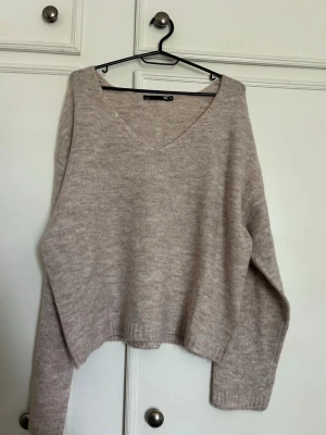 Beige stickad tröja med v-ringning - Mysig beige stickad tröja med djup v-ringning och avslappnad passform. Tröjan har långa ärmar och ribbade muddar vid ärmslut och nederkant. Perfekt för lager på lager och enkel att matcha till olika outfits.