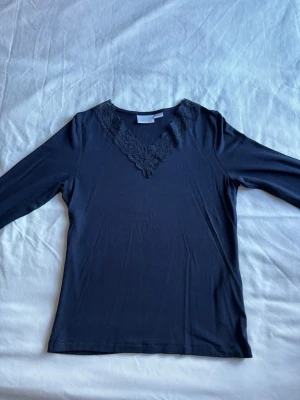 Svart långärmad topp med spets - Snygg svart långärmad topp med v-ringning och spetsdetalj runt halsen. Perfekt att styla till jeans eller kjol för en trendig look. Materialet känns mjukt och stretchigt, vilket gör den bekväm att bära hela dagen.