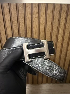 Svart bälte  - Snyggt svart bälte. Bältet har vit kontrastsöm längs kanterna och är gjort i skinn med en stilren och klassisk look. Perfekt accessoar för att lyfta din outfit.
