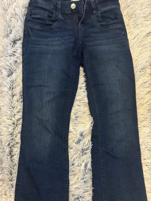 Mörkblå bootcut jeans - Säljer ett par mörkblå bootcut jeans i klassisk femficksmodell. Jeansen har en snygg tvätt med lätt slitning framtill och raka ben som går ut lite nedtill. Tillverkade i mjukt denimtyg med knapp och dragkedja i gylfen. Perfekta för en avslappnad och trendig look.
