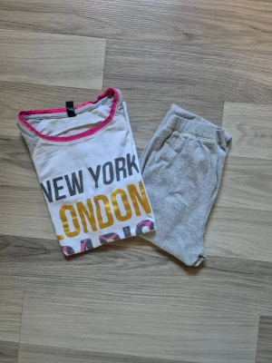 Pyjamas med tryck New York London Paris - Tvådelad pyjamas med t-shirt och byxor. T-shirten är vit med grå ärmar och har tryck i svart, gult och rosa med texten 'NEW YORK LONDON PARIS'. Rosa kant vid halsen. Byxorna är ljusgrå med resår i midjan. Perfekt för mysiga kvällar.