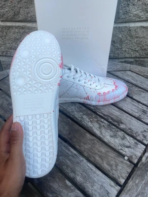 Skor - Unika vita sneakers med rosa färgstänk över hela skon. Tillverkade i slätt läder med klassisk låg siluett och vita snören. Snygg detalj med färgstänken som ger en kreativ och trendig vibe. Perfekta för dig som vill sticka ut.