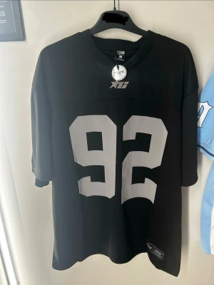Svart sporttröja med nummer 92 - Säljer en svart sporttröja med stora vita siffror 92 på framsidan. Tröjan har en lös passform, korta ärmar och V-ringad hals. Tillverkad i ett lätt och ventilerande material, perfekt för träning eller match. flera storlekar
