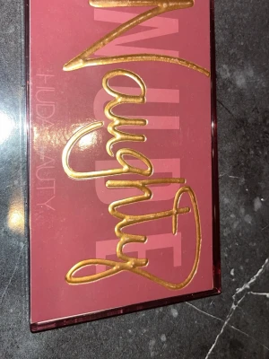 Naughty Nude ögonskuggspalett - Säljer en Huda Beauty Naughty Nude ögonskuggspalett med 18 nyanser i varma och kalla toner. Paletten innehåller matta, skimrande och metalliska skuggor i rosa, guld, brunt, orange och nude. Perfekt för kreativa och intensiva ögonlooks. Snygg design med guldig text på locket.