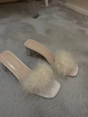 Vita pumps med fluff och klack - Säljer ett par vita pumps med transparent klack och fluffig, ljusbeige fuskpälsdetalj över foten. Skorna har öppen tå och fyrkantig framdel, vilket ger en trendig look. Perfekta för dig som vill sticka ut med en unik och lekfull stil.