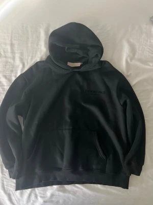 Svart Essentials Fear of God hoodie - Snygg svart hoodie från Essentials Fear of God med diskret logga på bröstet. Klassisk passform med stor huva och magficka. Tillverkad i mjukt bomullsmaterial som känns riktigt skönt mot huden. Perfekt för dig som gillar streetwear och enkel stil.