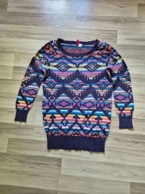 Flerfärgad stickad tröja med mönster - Stickad tröja i lila bas med färgglatt geometriskt mönster i blått, rosa, orange, gult och vitt. Tröjan har rund halsringning, långa ärmar och ribbade muddar vid ärmslut och nederkant. Perfekt för dig som gillar färg och unika plagg.