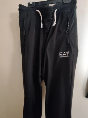 EA7 byxor - Snygga svarta mjukisbyxor från EA7 Emporio Armani med vita dragsnören i midjan och dragkedjor på fickorna. Byxorna har en avslappnad passform och EA7-logga tryckt på vänster ben. Perfekta för chill eller streetwear.