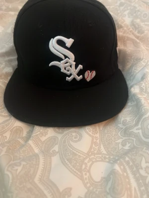 Oanvänd Svart White Sox keps med brodyr - Snygg svart keps med White Sox-logga i vitt framtill och ett litet hjärta med basebollmönster. På sidan finns ett broderat World Series-märke. Klassisk rak skärm och djup passform, perfekt för dig som gillar sportig streetstyle.
