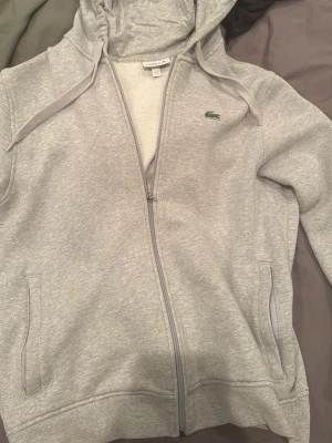Grå zip-hoodie från Lacoste XL - Snygg ljusgrå hoodie från Lacoste i klassisk passform med dragkedja, huva och två fickor. Ikoniska gröna krokodilloggan på bröstet. Mjuk insida och stilren design. Säljer pga fel storlek                                     Tveka inte på att höra av dig om frågor, bilder osv! Skickar snabbt!