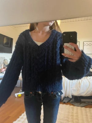 Mörkblå fluffig stickad tröja - Mysig mörkblå stickad tröja med v-ringning och breda ärmar. Tröjan har en fluffig struktur och ribbstickade muddar vid ärmslut och nederkant. Perfekt för kyliga dagar och enkel att matcha med jeans eller kjol.