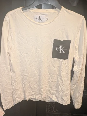 Creme färgad långärmad tröja från Calvin Klein - Creme färgad långärmad tröja från Calvin Klein Jeans med svart bröstficka och stor CK-logga. Tillverkad i mjuk bomull och har en klassisk rund halsringning. Inga synliga fläckar eller defekter. Skapa en bundle för bättre pris