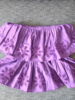 Ljuslila offshoulder topp med broderi - Säljer en söt ljuslila offshoulder topp med volang och broderade blommor. Toppen har elastisk smock i ryggen och är ärmlös, perfekt för sommarens varma dagar. Materialet känns som bomull och toppen har en luftig och romantisk vibe.