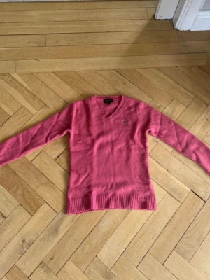 Rosa stickad tröja från GANT - Säljer en rosa stickad tröja från GANT i storlek XXL som motsvarar 158-164. Tröjan har V-ringning, ribbade muddar och en broderad GANT-logga på bröstet. Perfekt för dig som gillar klassisk och stilren design. Materialet är mjukt och skönt.