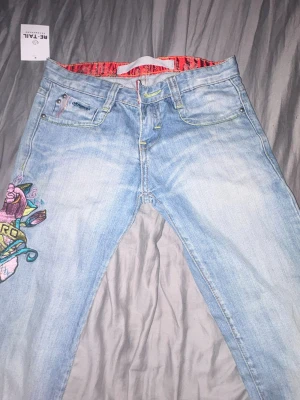 Lågmidjade Ljusblå bootcut jeans med broderad detalj - Unika ljusblå jeans med färgglad broderad design på ena benet, där motivet inkluderar en ros, fågel och texten 'ZERO'. Jeansen har klassisk fyrficksmodell och raka ben. Materialet är denim i bomull och insidan har en röd mönstrad kant. Säljer då jag köpte fel strl helt oanvända!❤️