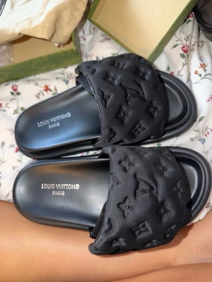 Svarta  sandaler - Stilrena svarta sandaler med bred vadderad rem och ikoniskt monogrammönster. Mjuka och bekväma med en lyxig känsla, perfekta för en trendig look. Tillverkade i textil och syntetmaterial.