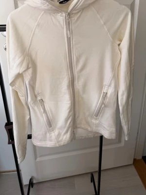 Vit hoodie med dragkedja och huva - Säljer en stilren vit hoodie med dragkedja framtill och två fickor med dragkedja. Hoodien har långa ärmar och en klassisk huva. Perfekt för en avslappnad look och enkel att matcha med andra plagg.