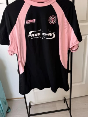 Svart och rosa sporttröja Free Spirit - Snygg svart sporttröja med rosa detaljer och tryck fram och bak. På bröstet står det 'FREE SPIRIT' och 'Coolane', samt ett märke med text. Baksidan har stort 'USA'-tryck och texten 'Dream Design Department'. Perfekt för träning eller streetwear.