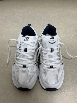 Vita New Balance sneakers - Snygga vita sneakers från New Balance med meshpaneler och detaljer i silver och svart. Skorna har en klassisk låg siluett, vita snören och en bekväm sula. Perfekta för dig som gillar sportig och clean stil.