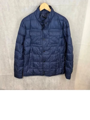 Mörkblå pufferjacka med fickor - Snygg mörkblå pufferjacka med quiltad design och hög krage. Jackan har två bröstfickor med lock och knappar framtill. Perfekt för kalla dagar och ger en stilren look. Tillverkad i ett glansigt material som håller värmen bra.
