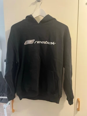 Svart Reebok hoodie med logga - Svart hoodie från Reebok med stor vit logga på bröstet. Klassisk känguruficka framtill och huva med grått innerfoder. Perfekt för en avslappnad och sportig stil. Mjuk och skön bomullsblandning, ribbade muddar vid ärmar och nederkant.