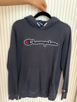 Mörkgrå Champion hoodie med logga - Snygg mörkgrå hoodie från Champion med stor broderad logga på bröstet och klassisk känguruficka. Tröjan har huva och ribbade muddar vid ärmslut och nederkant. Perfekt för en avslappnad streetstyle-look.