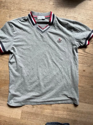 Äkta Moncler t-shirt - Snygg grå t-shirt från Moncler med v-ringad krage och röd, vit och marinblå rand på krage och ärmslut. Klassisk Moncler-logga på bröstet. Funkar med precis alla stilar därför unisex men jag skulle tipsa till män framförallt. Väldigt stilig. Den är äkta för den går att scanna!!!
