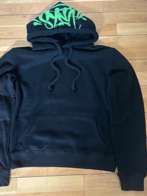 Svart hoodie med grön graffiti-huva - Säljer en svart hoodie med stor ficka framtill och huva med grönt graffiti-tryck. Hoodien har tjocka snören och ribbade muddar vid ärmslut och midja. Perfekt för dig som gillar streetwear och vill sticka ut med en unik detalj på huvan.