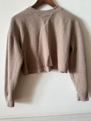 Beige croppad stickad tröja - Säljer en beige croppad stickad tröja med långa ärmar och ribbade muddar. Tröjan har en avslappnad passform och rundad halsringning med ribbdetalj. Perfekt för lager på lager och enkel att styla till olika outfits.